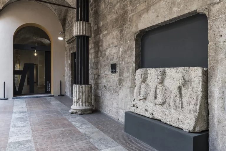 Ingresso alla sezione romana dal cortile interno. In primo piano un monumento funerario romano in pietra con i busti-ritratto di tre defunti.